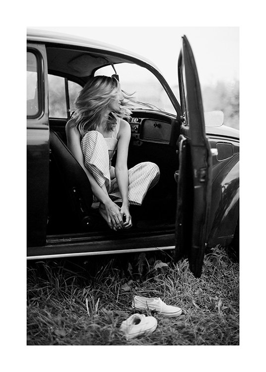  – Schwarz-Weiß-Fotografie einer Frau in einem Oldtimer mit offener Tür. Ihre Schuhe liegen vor dem Auto.