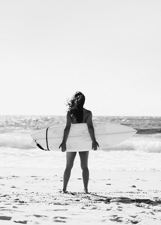  – Schwarz-weiß-Fotografie von einer jungen Frau, die ihr Surfbrett hinter sich hält, im Hintergrund das Meer