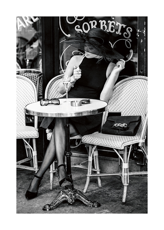  – Schwarz-weiß-Fotografie einer Frau, die draußen vor einem Café sitzt, einen Hut trägt und eine Zigarette anzündet