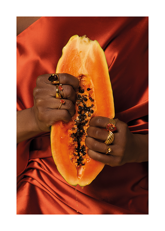  – Frau in einem Satinkleid, die goldene Ringe trägt und ihre Finger in eine Papaya drückt