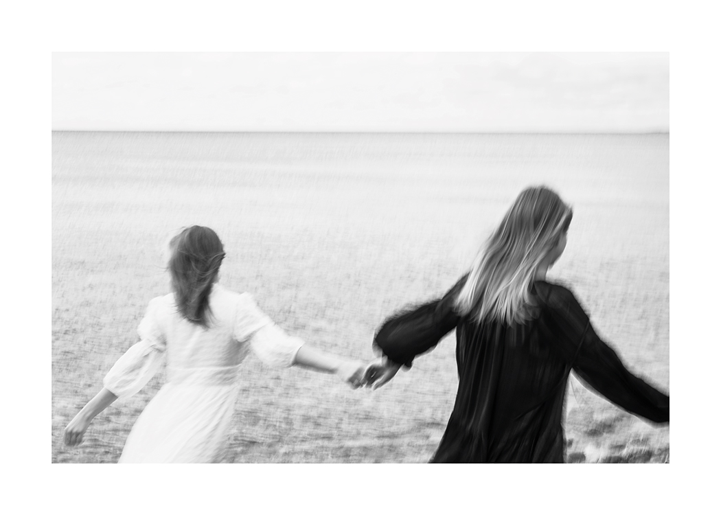  – Schwarz-weiß-Fotografie von zwei Frauen, die einander an den Händen halten und über ein Feld laufen